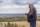 Une personne aux longs cheveux gris se tient à l'extérieur, avec un paysage vallonné et herbeux et un ciel nuageux au loin.